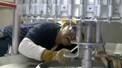 Screen shot of video with man with protective equipment on, holding a wrench, looking up into machine